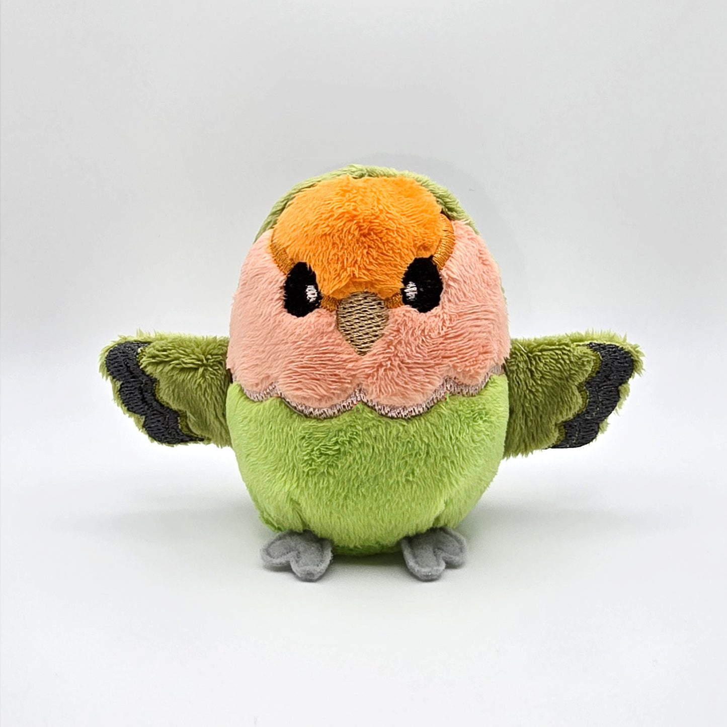 Round orange, peach and green plush lovebird with wings outspread. Face and features embroidered. Front view.