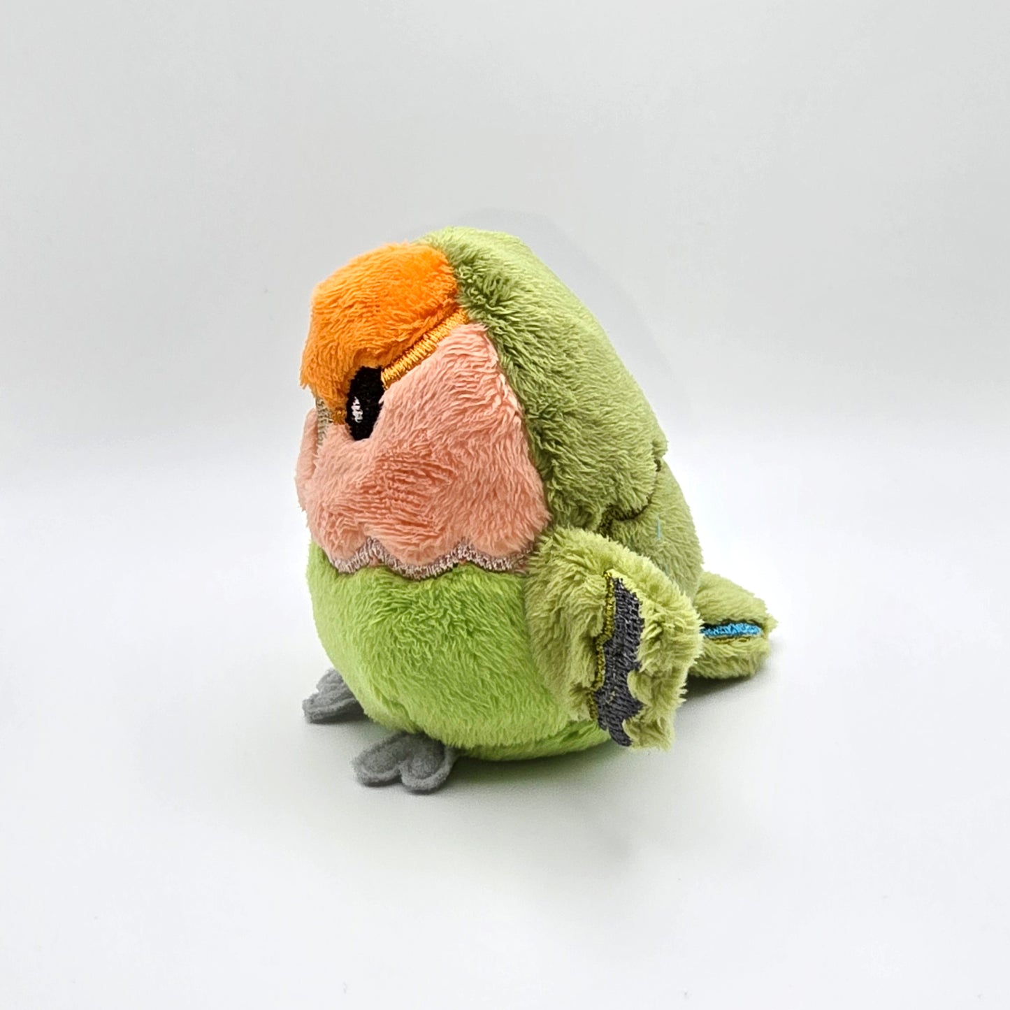 Round orange, peach and green plush lovebird with wings outspread. Face and features embroidered. Side view.