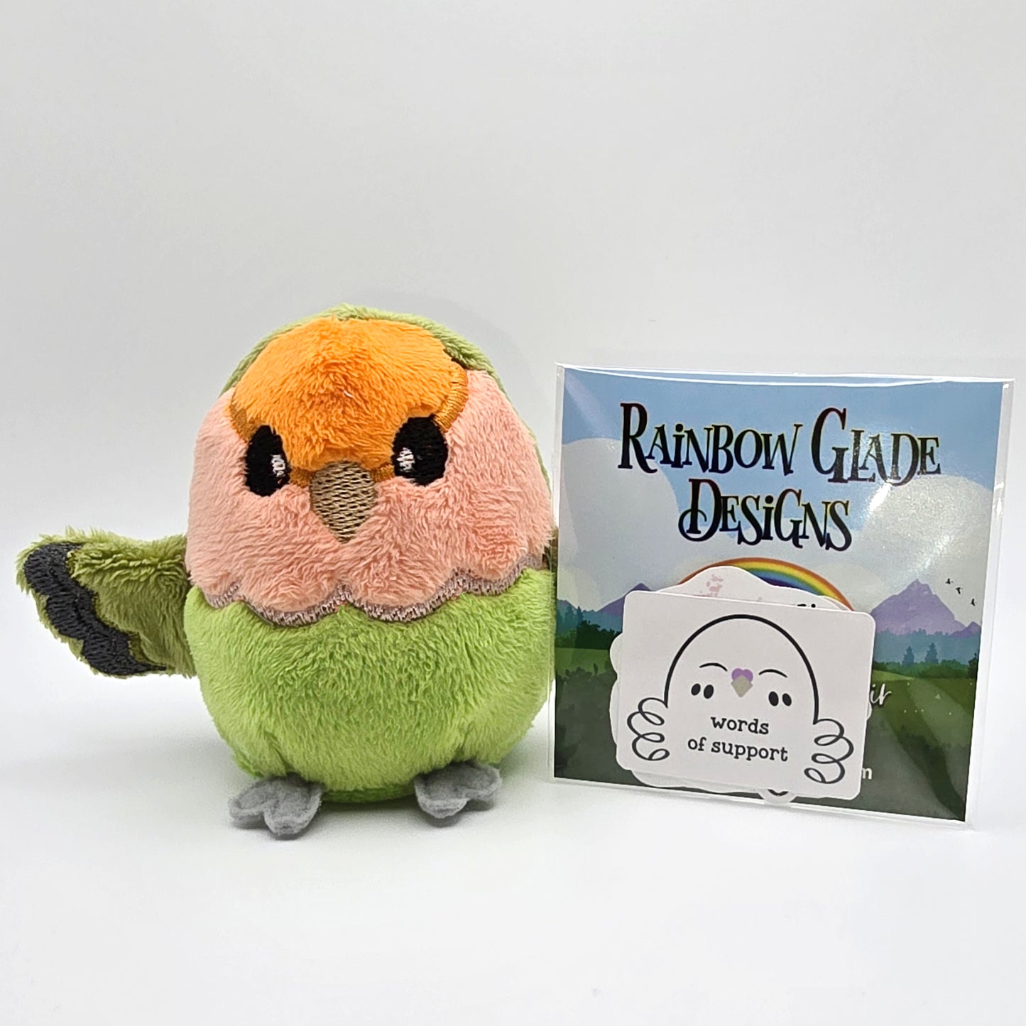 Round orange, peach and green plush lovebird with wings outspread. Face and features embroidered. Front view showing accompanying sticker set.