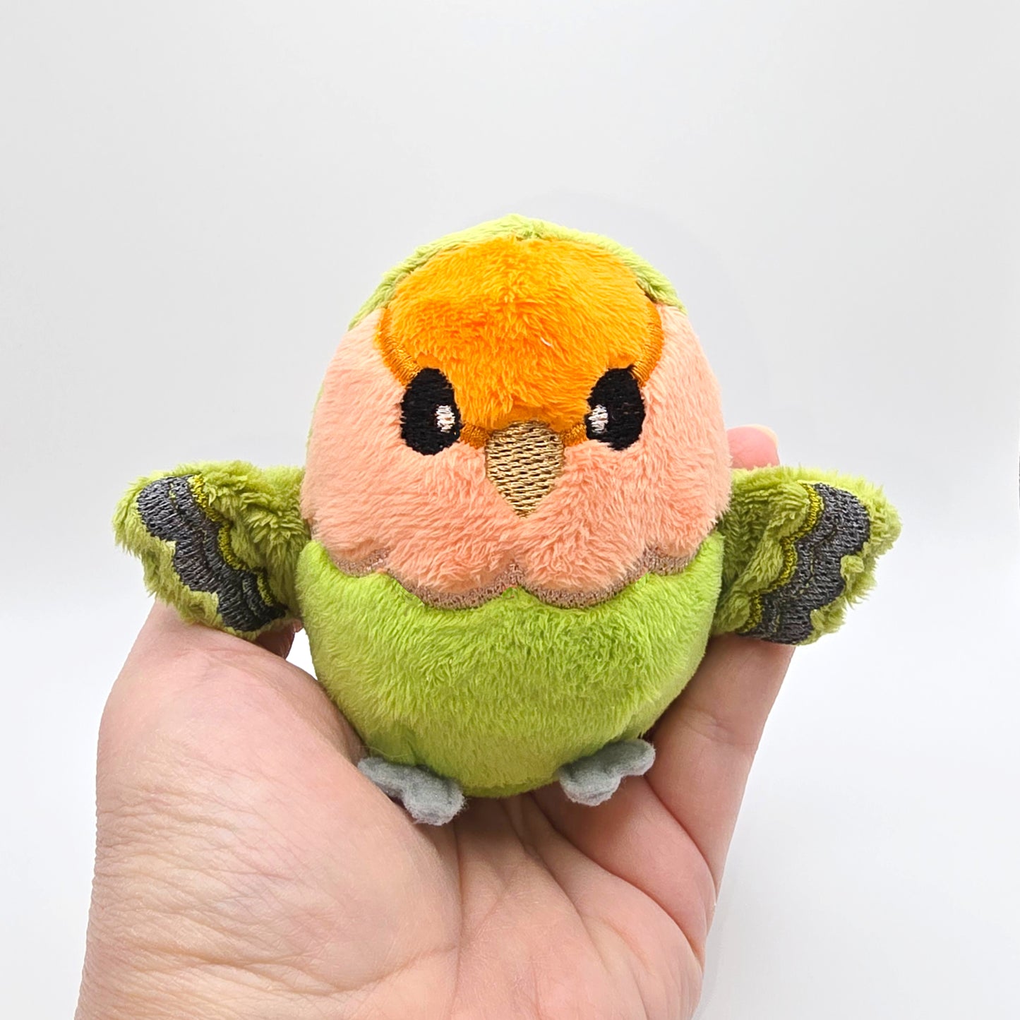 Hand holding round orange, peach and green plush lovebird with wings outspread. Face and features embroidered. Front view.